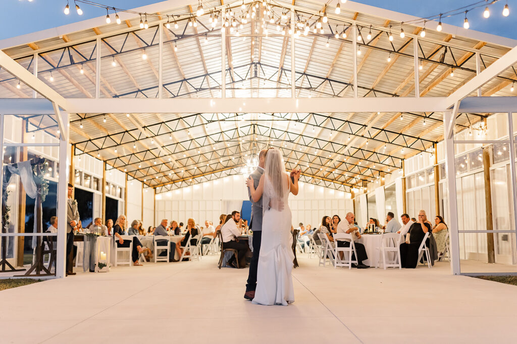 Bride and groom dance under the string lights on the outdoor dance floor.  Wedding reception barn has greenhouse vibe with clear enclosure.  Seated guests enjoy the wedding reception.