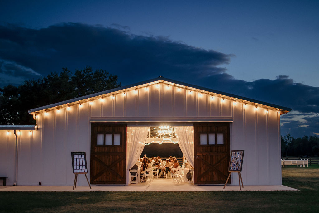 Evening photo of the wedding reception barn framed in string lights and the large stained wood barn doors are standing open to show a happy wedding reception taking place.  The sun is setting and casting a shadowed glow over the barn.