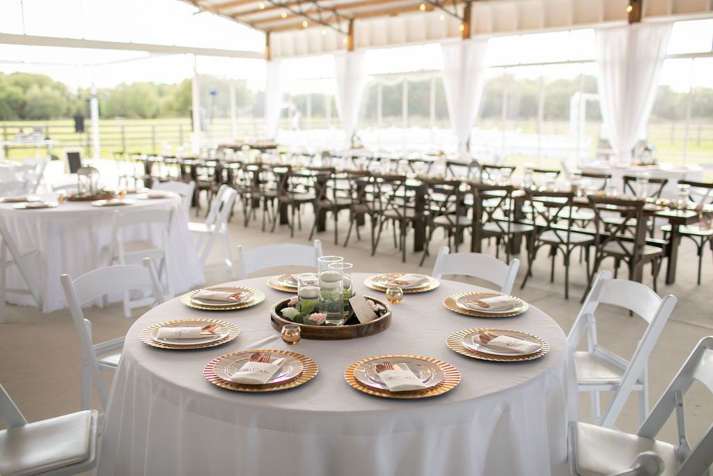 Wedding reception space set up for 100 guests.  Farm style and round tables with white linens and gold chargers. Clear greenhouse style enclosure provide open views of farm.