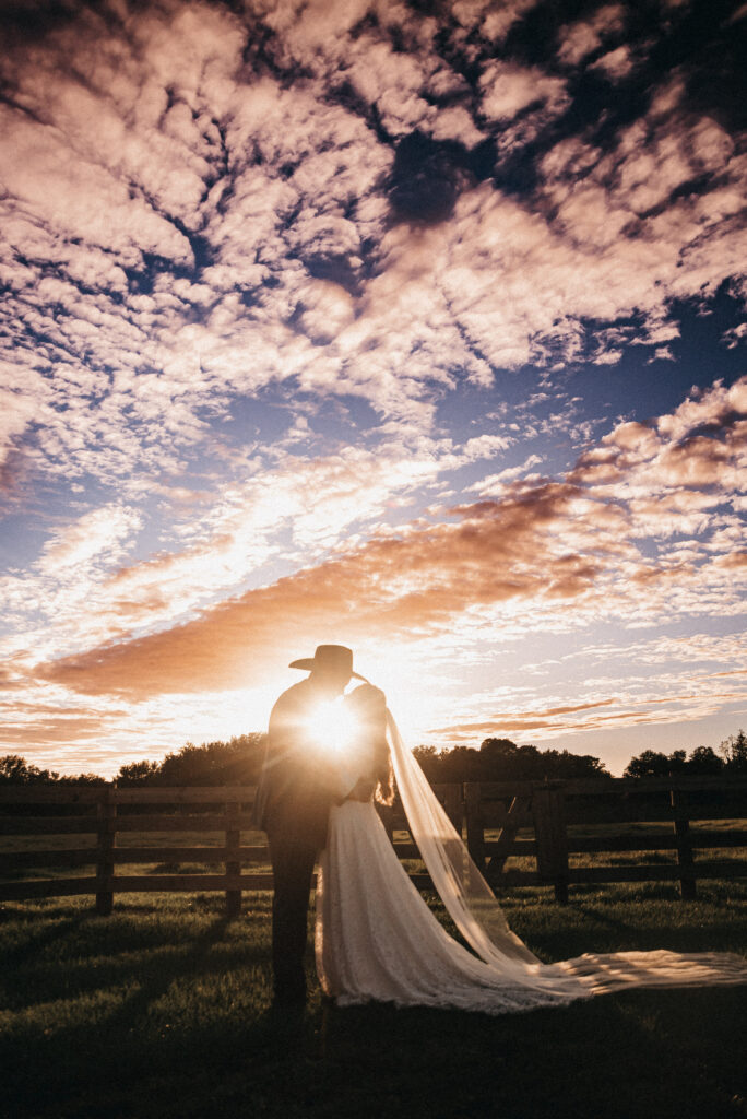 Bride and groom share a kiss while the sun casts a bright glow in the center of their embrace.  The blue sky with puffy clouds provide an amazing sunset backdrop.