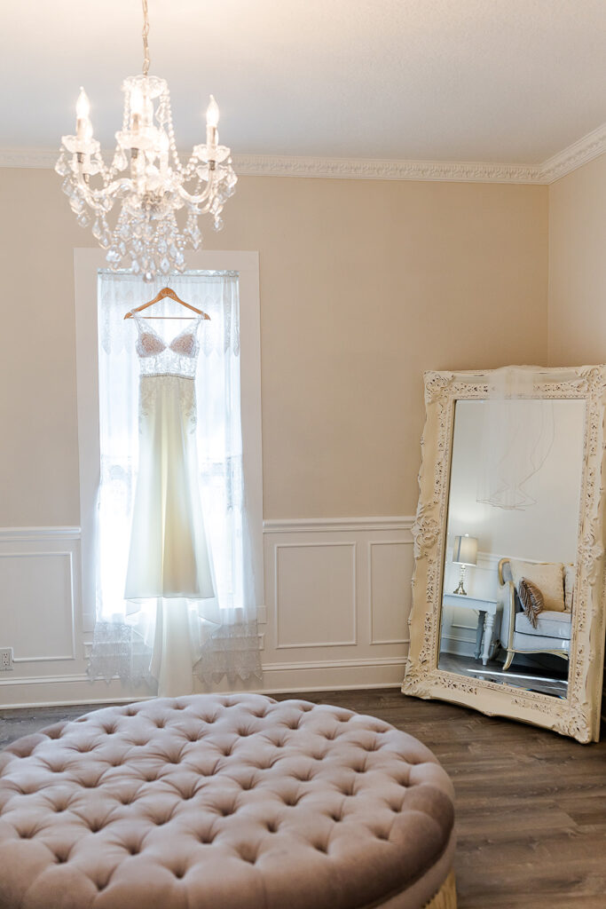 Bridal suite with vintage vibe.  Wedding dress hangs in light filled window under the crystal chandelier while ornate white framed tall leaning mirror reflects the room and bougie furnishings.