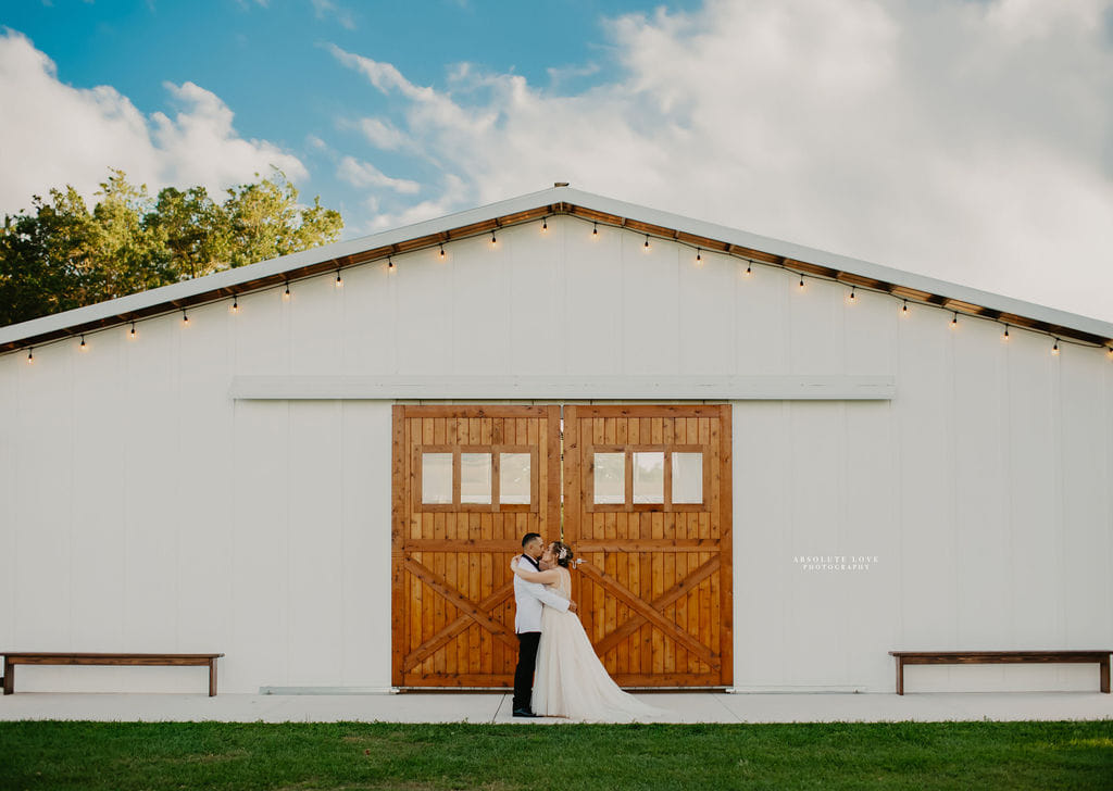 Young bride and groom in front of white wedding Big Barn Pavilion.  Stained wood doors for backdrop and a blue sky with puffy clouds. String lights frame the barn for added charm.