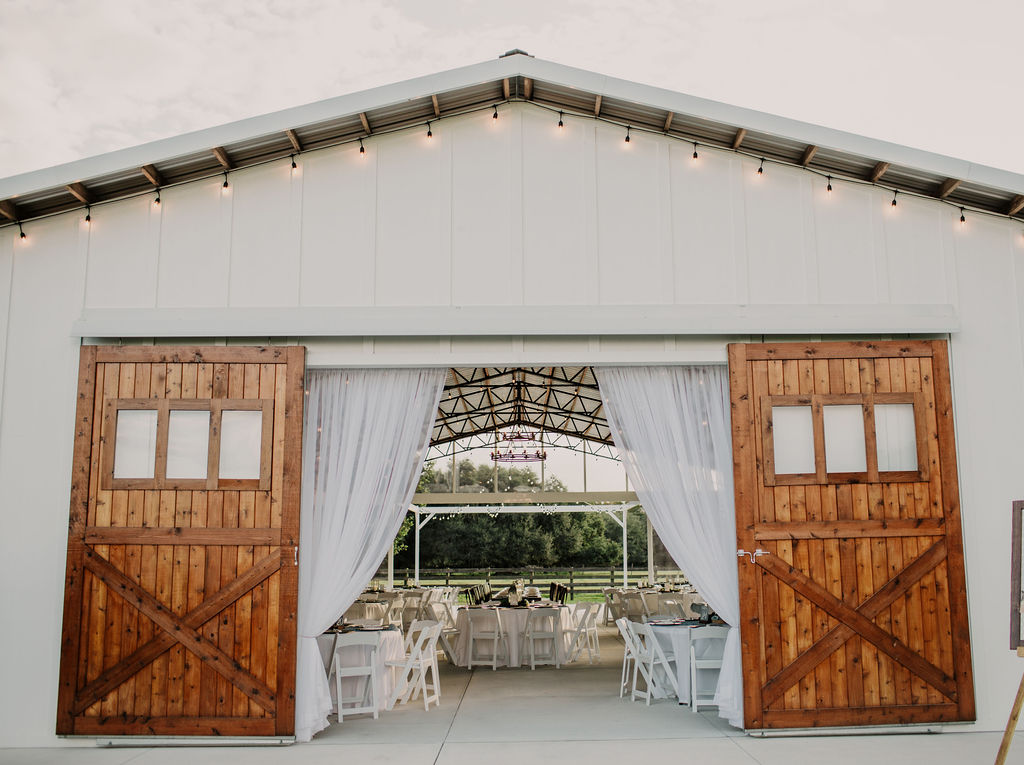 Front of big white barn with large stained barn doors. The lighted string lights are lining the eave of the barn. Barn doors are open to the view of the open area at the outside dance floor. Tables and chairs can be seen on a beautiful day.