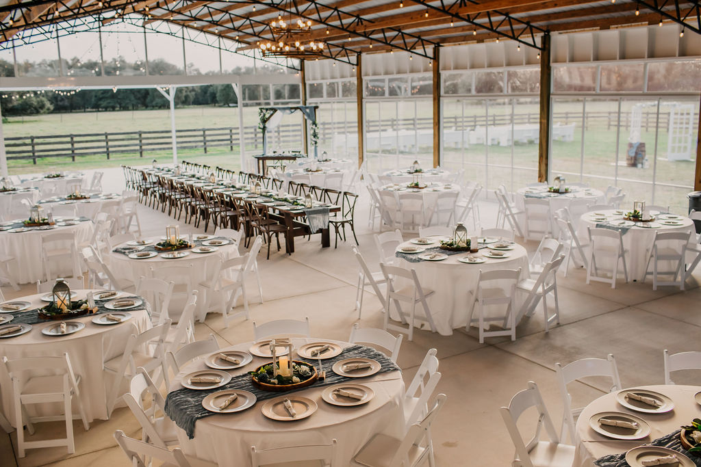 The Heart D Farm Wedding & Event Venue big barn wedding reception pavilion. Greenhouse vibe in a large open space with clear moving enclosure for unobstructed views of the green pastures and board fencing. The space is set up for 175 guests with the sweetheart table and decorated wedding arch in the background. Just outside the big barn pavilion is the large outdoor dance floor. Inside are four wood farm tables lined up for 32 seated guests with wood cross-back chairs. The other tables are round with white table cloths and white garden chairs with silver chargers, wood round centerpieces, lighted candles and dusty blue table runners. Outside you can see the ceremony site with white church pews and our decorated French door aisle entry with whisky barrels and large lanterns.