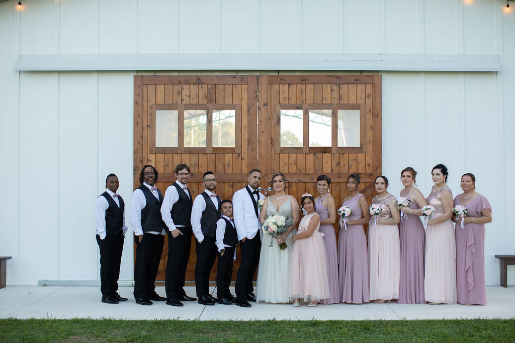 Bride and groom with wedding party stand in front of the white barn with large stained barn doors. The groom and groomsmen are sharply dressed in black and white while the bridesmaids are dressed in soft pinks and lavenders.
