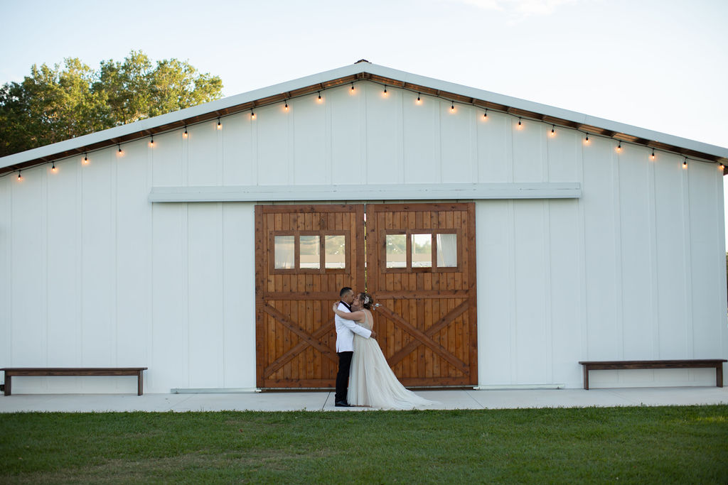 Sweet couple kiss in an embrace in front of the enormous stained wood barn door on the front of the Big Barn wedding reception building. The bride is dressed in a flowing off white filmy wedding dress and the groom is looking sharp in his black slacks and white blazer with a black lapel. The barn is white and the stained wood doors are a beautiful contrast to the white background. Market lights line the eave of the barn as the sun is beginning to cast its golden glow.