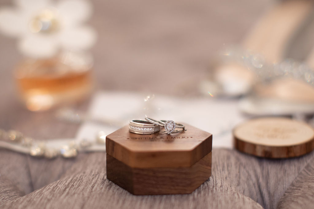 His and hers wedding bands and diamond ring on beautiful small wood box with a blurred background of perfume bottle, crystal necklace and wedding heels. All set on a tufted gray surface.
