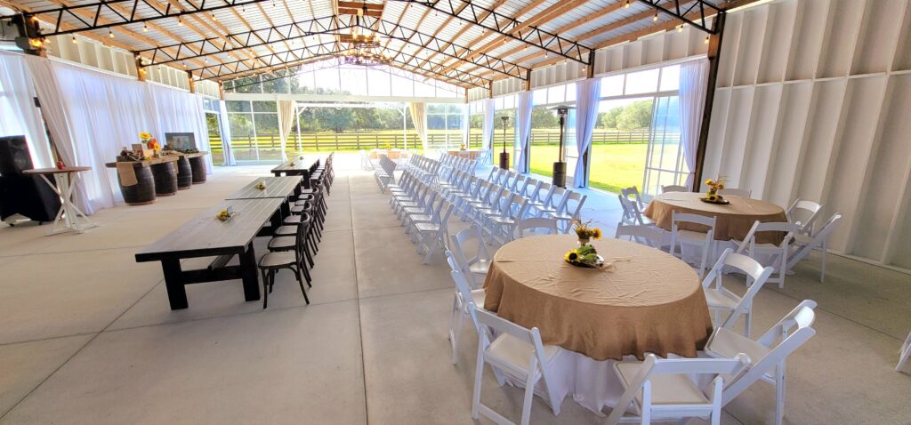 Big Barn Pavilion with rowed seating of white chairs and a row of brown cross back chairs at the long wooden farm tables set for the family.  There are a few round tables with white and burlap table cloths and white garden style chairs.  The barn has a clear enclosure that allows for sunny views of the green pasture and four board fence.