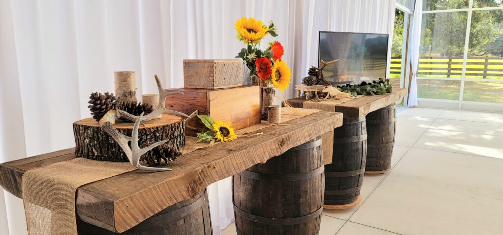 Four brown whiskey barrels are in line with rustic wood tops.  Displayed on top are wood boxes, a large round wood tree chunk with bark still on the outside.  There is a set of deer horns, pine cones and sunflowers.  There is a TV with greenery around it.  There is a white draped background and also a clear view of the four board fence, trees and pasture.  