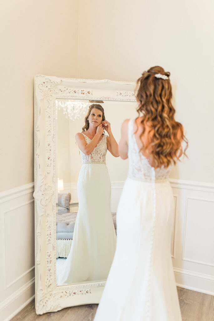 Lovely bride fixes her right earring while looking into the large ornate white mirror in the bridal suite. Her white wedding dress is sleeveless with lace top and solid white lower half and is fitted. Her long sandy blonde hair cascades down her back on the open area of her dress. The bridal suite is painted in soft whites.