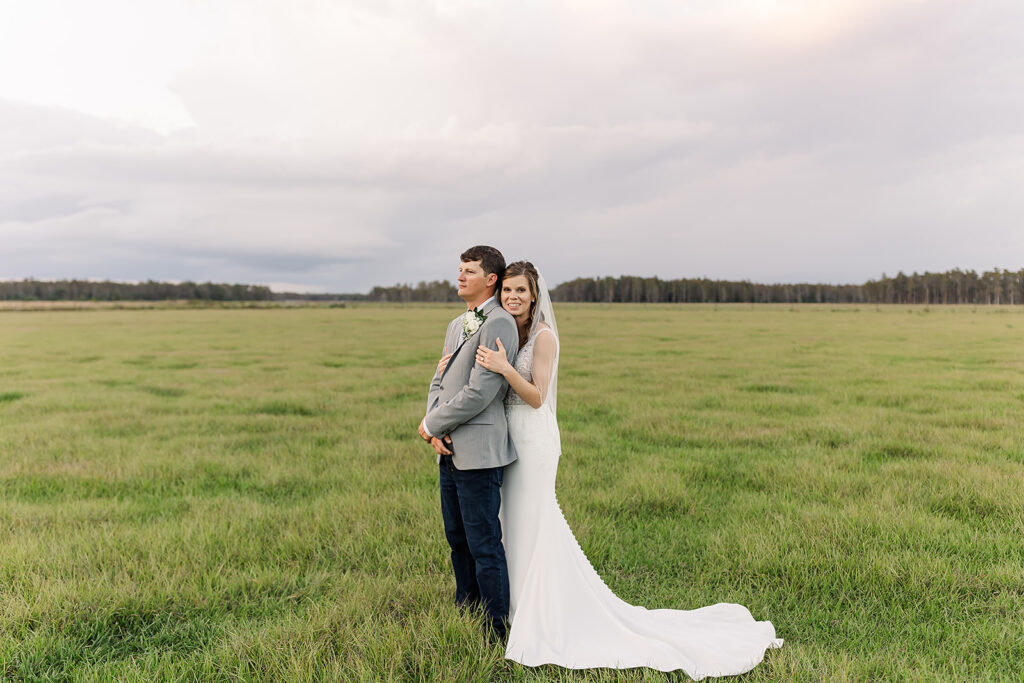 Newly wedded couple stand in an open pasture with a long distant view behind them of only grass and in the distance stands of cypress trees. The groom is facing left and the bride is standing behind him holding his arms and gently laying her cheek on his shoulder. He has his hands folded in front of him and is gazing into the distance as if looking to their future. He is wearing a gray jacket with white and blue boutonniere and dark blue jeans. The bride is wearing a fitted white gown with a solid white train and waist-length bridal veil pinned to her blonde hair. The sky is large and somewhat overcast.