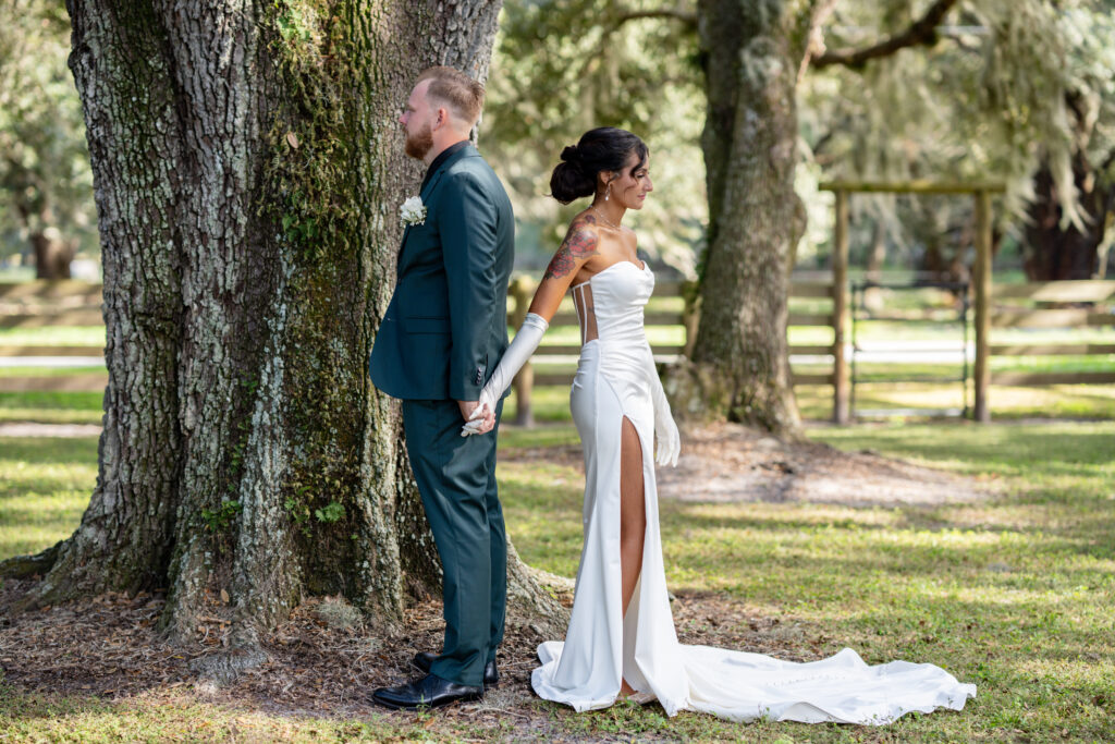 Bride and groom standing under large oak trees with more oak trees and a four board fence in the background. Couple are posing for their first look standing with their backs to each other and holding his left and her right hand together. He is wearing a dark green suit with black shoes and she is stunning in her fitted wedding dress with solid white train displayed around her feet. The strapless white wedding dress has a slit to her upper right thigh and bares her shoulders and back. The long white wedding gloves are the perfect detail for her look.