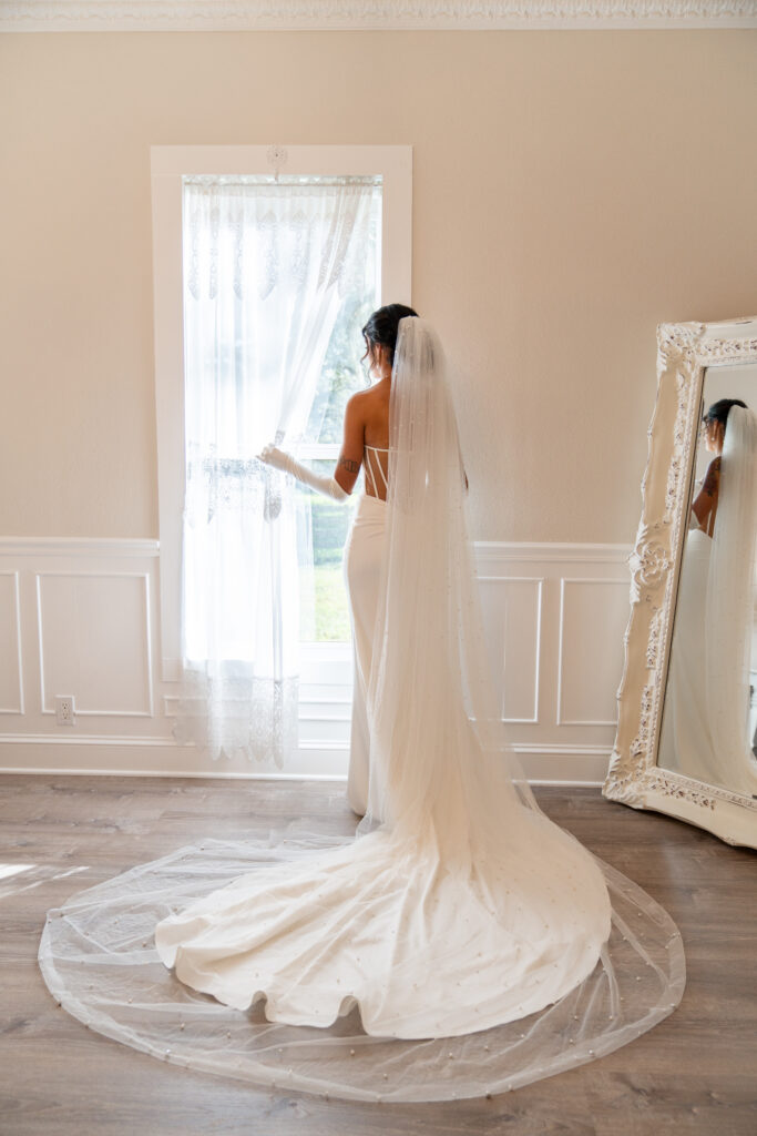 A lovely bride in a fitted strapless, backless white wedding dress stands at the window with the lace curtain drawn back to look upon her day. She has a cathedral veil pinned to her dark hair and it cascades over and around her train in a circular shape on the floor. The vintage styled bridal suite is done in soft whites and grays with wains coating white trim. In the corner stands a gorgeous full length leaning white mirror with an ornate frame and the bride's reflection. It is a beautiful photo.