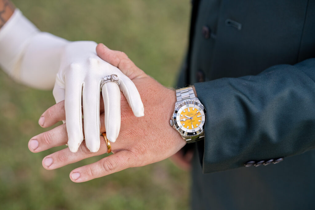 Bride and groom displaying their hands. Hers with a long white glove and the wedding band and diamond ring on her ring finger. His with a nice watcha and gold wedding band in an outdoor ceremony setting.