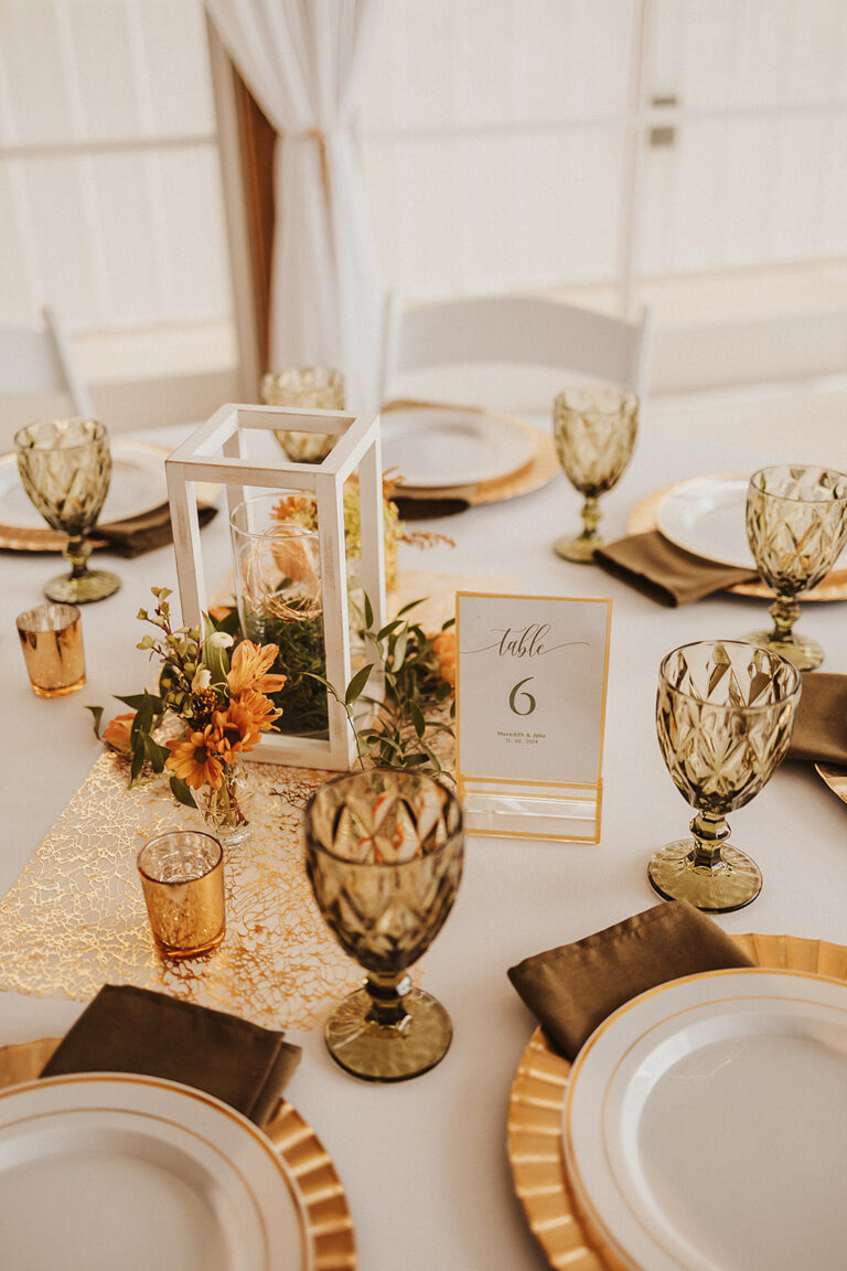 Gorgeous table setting with gold chargers, olive green water goblets, green cloth napkins, soft orange wild flowers and white lantern centerpiece. Set and ready for the wedding guests to arrive.