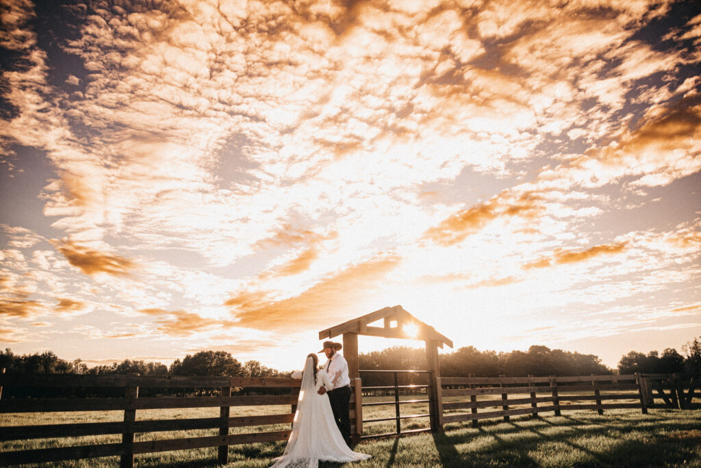 Newly wedded couple stand facing each other against a four board fence with an enormous brightly filled cloudy sky overhead. She is in a long white flowing wedding dress and a cathedral length wedding veil drawn out behind her and pinned in her long dark hair. The groom is wearing dark brown slacks, a white dress shirt and dark tie. He is sporting a brown cowboy hat which is perfect for the ranch style setting and vibe. The groom is taller than the bride and is romantically gazing downward into her eyes.
