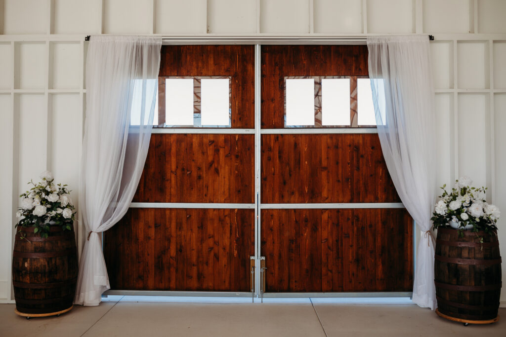 Interior photo the large stained barn doors with wood whisky barrels on each side and filmy white drapes framing the large doorway. White walls create the rustic background of the photo.