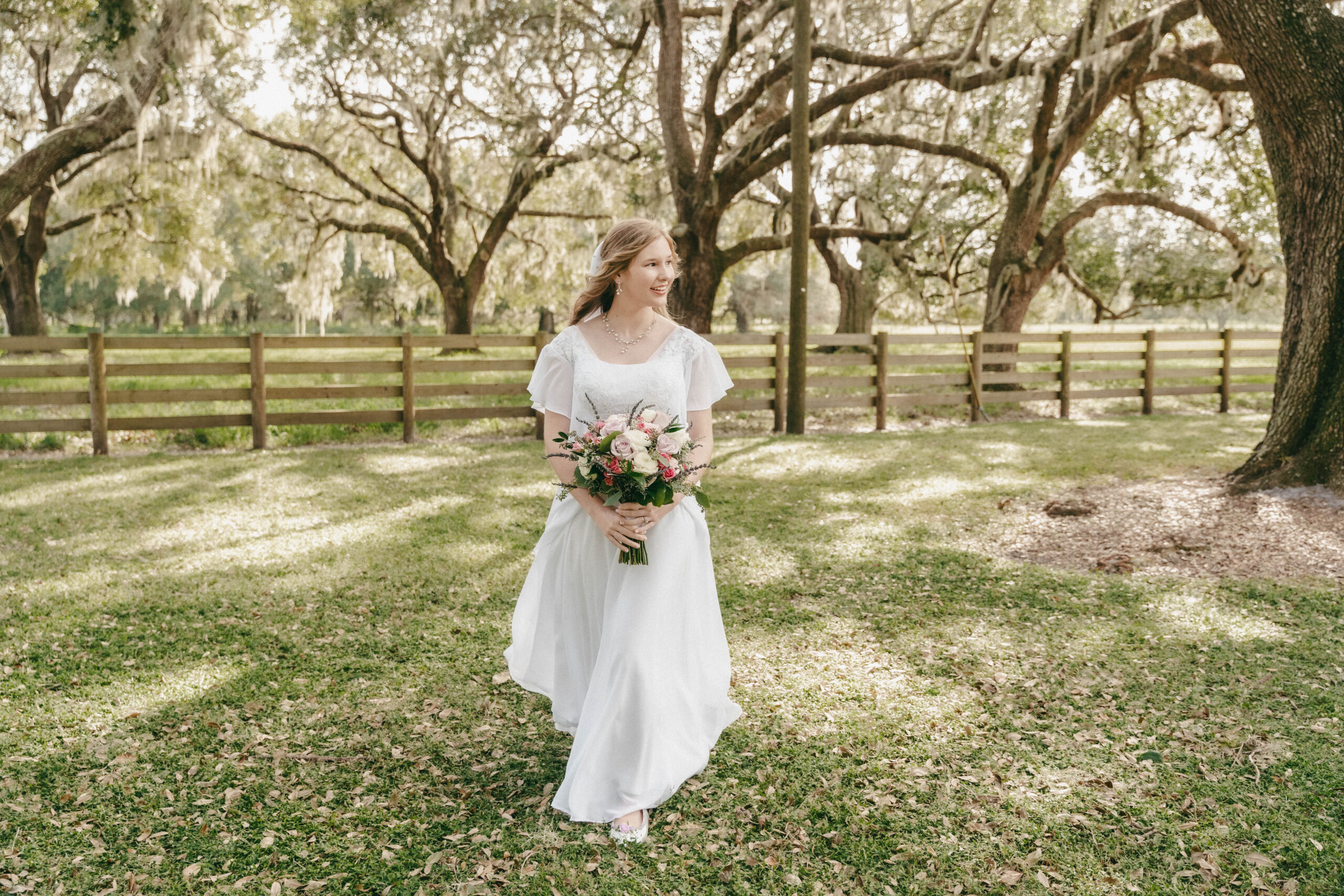 Lovely young bride on her wedding day before the outdoor ceremony. She is under the large oak trees with the sun filtering through. She is wearing a filmy white dress and carrying a colorful bouquet of flowers.