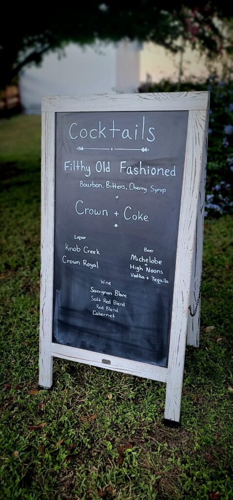This is a sign that set by the outdoor bar at this event. It reads "Cocktails, Filthy Old Fashioned, Bourbon, Bitters, Cherry Syrup. Crown & Coke, Liquor, Knob Creek, Crown Royal. Beer: Michelobe, High Noons, Vodka & Tequila. Wine: Sauvignon Blanc, Soft Red Blend, Red Blend, Cabernet