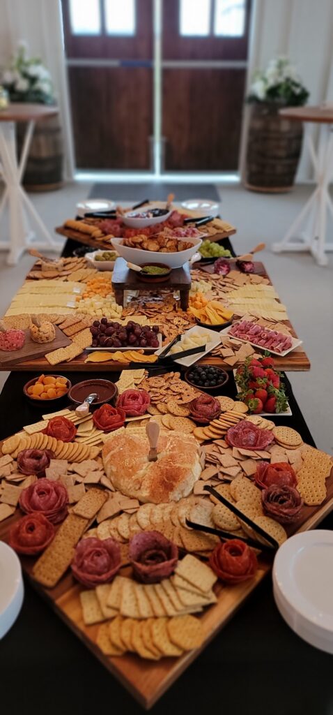 Long table with a black table cloth with grazing boards on top and the entire table and boards are filled with cheeses, grapes, sliced meats rolled into shapes like flowers, assorted crackers, smoked cream cheeses, pesto, cheese balls, strawberries, figs and green and black olives. It is colorful and very tasty in appearance.