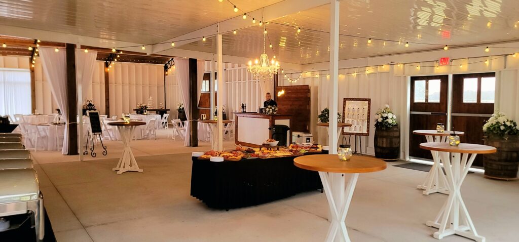 Cocktail area at the Heart D Farm Wedding Venue. The walls are white with string lights and a chandelier draped from the ceiling. Six high top farm style cocktail tables are placed around the room with an 8 ft food table with black floor length table cloth in the center filled with a grazing table for expected guests. The wood stained barn doors accent the white interior walls with a wooden whiskey barrel on each side. The custom bar is set and ready for guests. In the background you can see the reception area set with round tables draped in white linens with white chairs. Just moments away from the guests arriving.