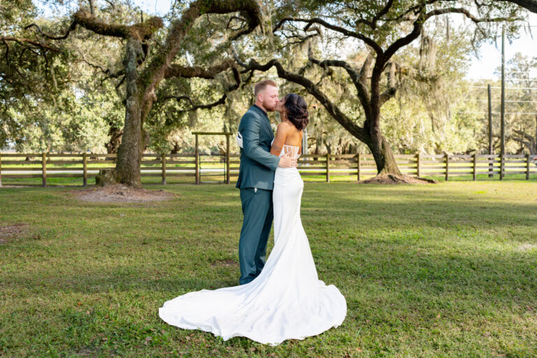 Beautiful couple embrace under the large ancient Live Oak trees on a green lawn with a four board fence in the background. He is wearing a dark green suit and her wedding dress is backless and white and very fitted with her train on the ground in a circle near their feet. She is also wearing long white gloves. Her hair is dark and done in a lovely updo. It is a very romantic scene.