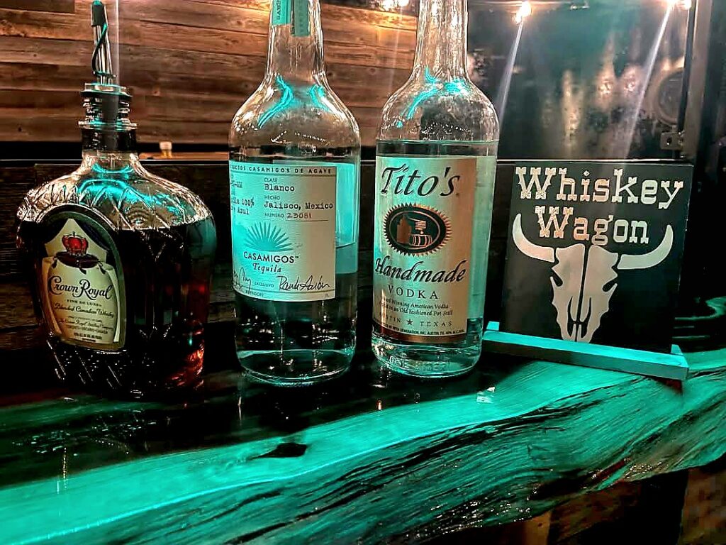 A shadowy closeup photo of a rustic wooden bar top with a bottle of Crown Royal Whiskey, Casamigos Tequila and Tito's Vodka with a small sign that says WHISKEY WAGON and a cow skull.
