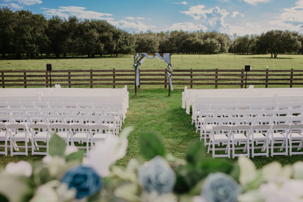 Outdoor ceremony site with white church pews and white garden chairs set up and awaiting guests. Everyone will over look the open field of the south pasture while the bride and groom stand in front of the traditional wedding arch decorated with dusty blue tulle and flowers.