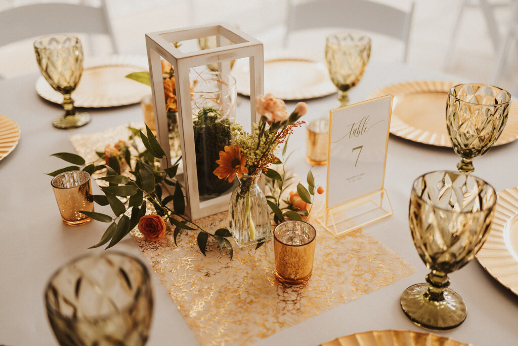 Gorgeous table setting on round table with white linen, white center lantern, gold chargers, wild flowers and olive green goblets at table number 7.