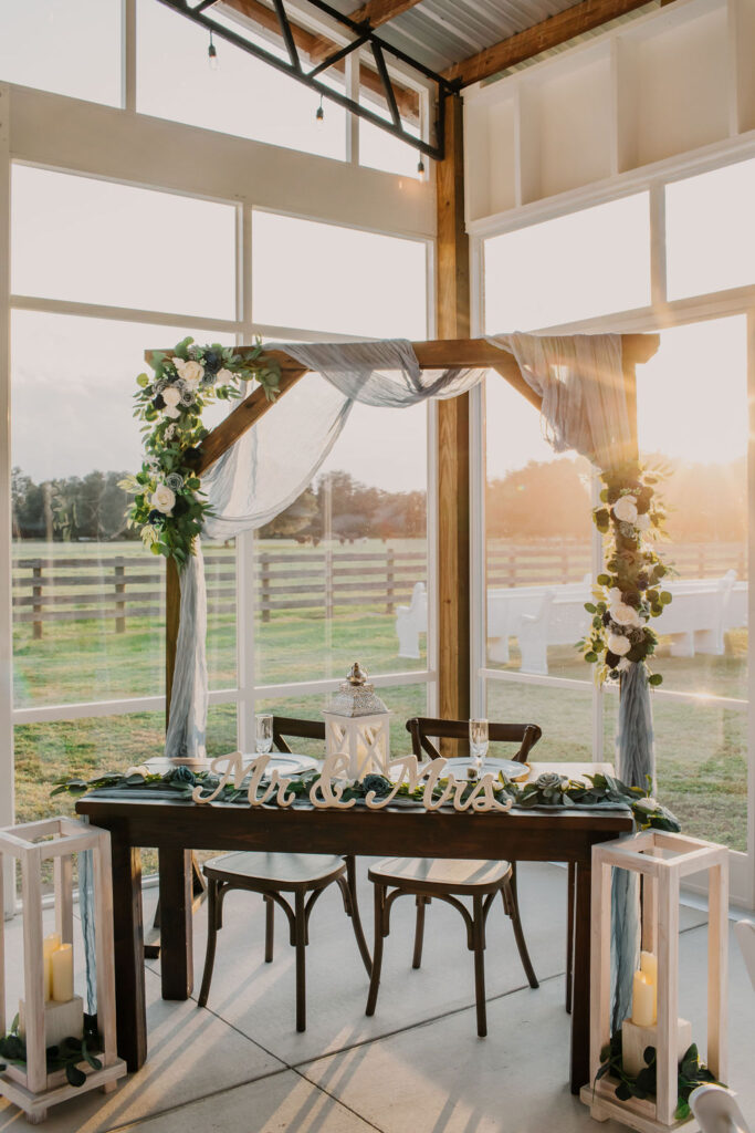 Sweetheart table decorated for the newly wedded couple.  The wedding arch is set behind the table and decorated in soft whites and blues.  The clear walls of the Big Barn Pavilion show the view of the white church pews with the sun setting and the pasture and cows in the background