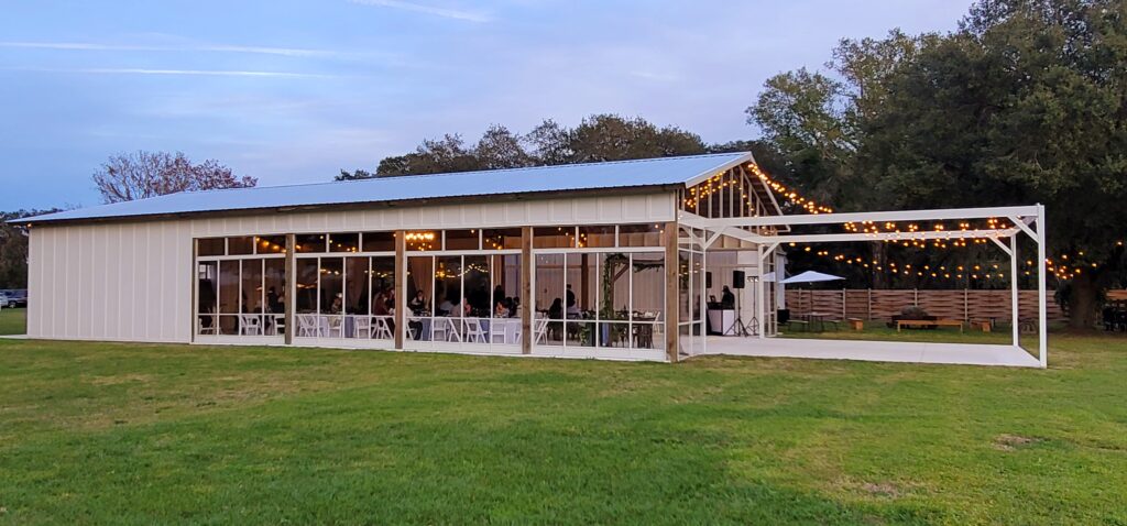 View of the white and clear glass Big Barn Pavilion from outside.  The sun is beginning to set and the string lights are transforming the look of the barn and the outdoor dance floor.  