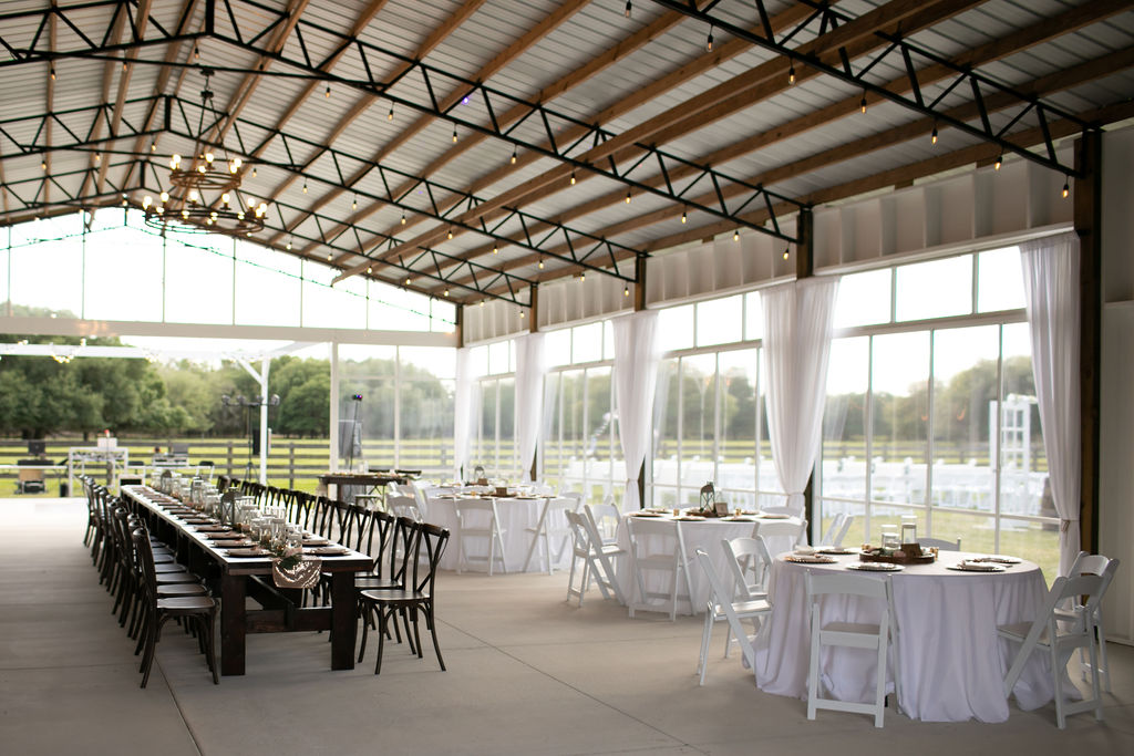 Interior of Big Barn Pavilion with white drapes tied back to show the view of the farm.  Long wood farm table and round white table are set and ready for the wedding guests to enjoy.  