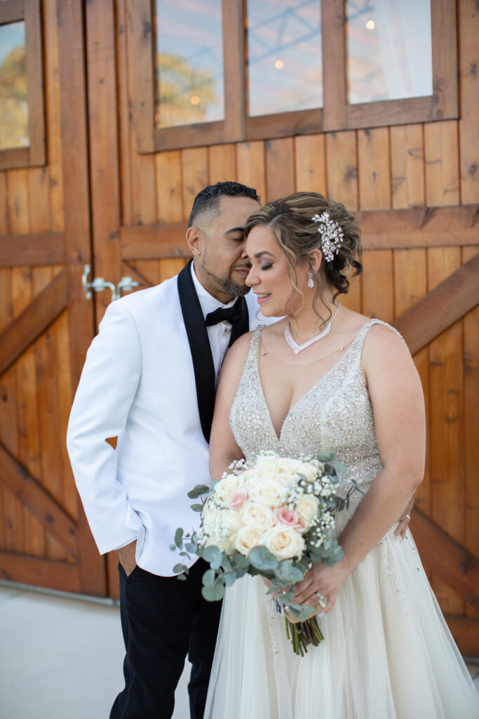 Newly wedded couple in front of the barn doors at the Heart D Farm Wedding Venue after their outdoor ceremony. He is leaning into her for a sweet moment. Both have their eyes closed