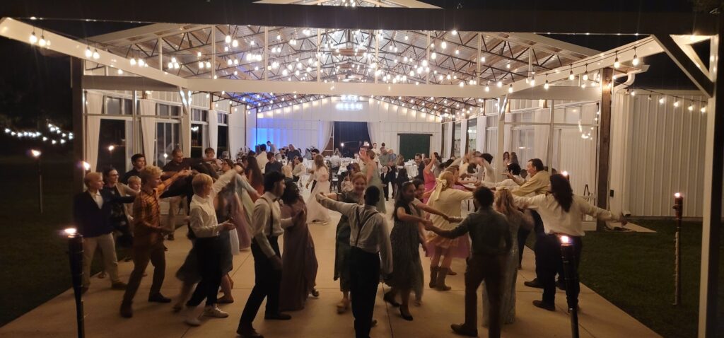 Dozens of wedding guests dance under the stars and the string lights on the outdoor dance floor after a wedding ceremony. Lots of fun being had by all.