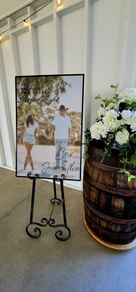 A poster sign of the couple walking hand in hand and dressed casually.  The sign is on an easel setting beside a whiskey barrel with soft white wedding flowers.