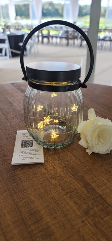 A glass lantern with fairy lights adorns a high top cocktail table with a single white rose and special QR code for the guests to upload their photos to the bride and groom.