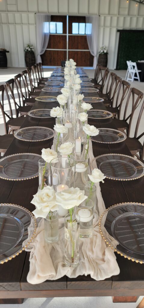 Simply elegant head table is set for the wedding guests at the Heart D Farm wedding venue in St. Cloud, FL.  The wood farm table is inside the Big Barn Pavilion and is set with simple white candles and dozens of single white roses each in a single clear bud vase.  The chargers are clear with gold beaded rim.  The huge stained wood barn doors create a lovely backdrop.