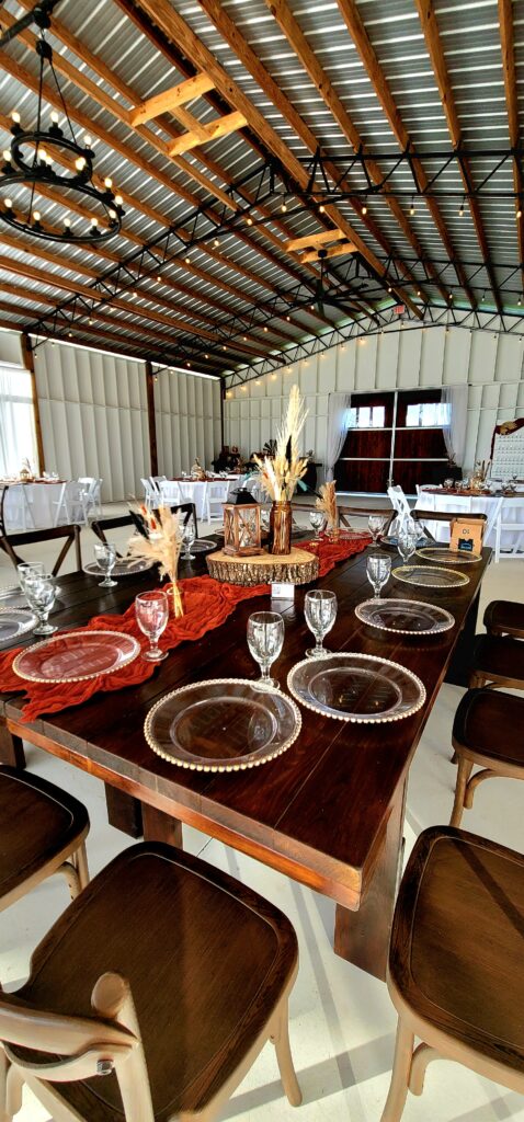 Inside our Big Barn Pavilion two of the wood farm tables are set side by side and are referred to as Kings Table.  Clear chargers with gold beaded rims, clear water goblets and a bit of western boho wedding decor adorn the tables.  All the wedding decor is included in the rental package