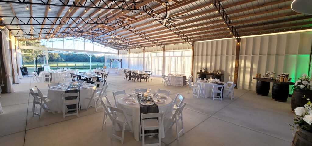 Big Barn Pavilion wedding reception area with clean enclosure open to the view of the green pastures.  The white drapes are closed to filter the setting sun.  The round tables are draped with floor length white linens.  Ready for the wedding guests to arrive after the outdoor ceremony.