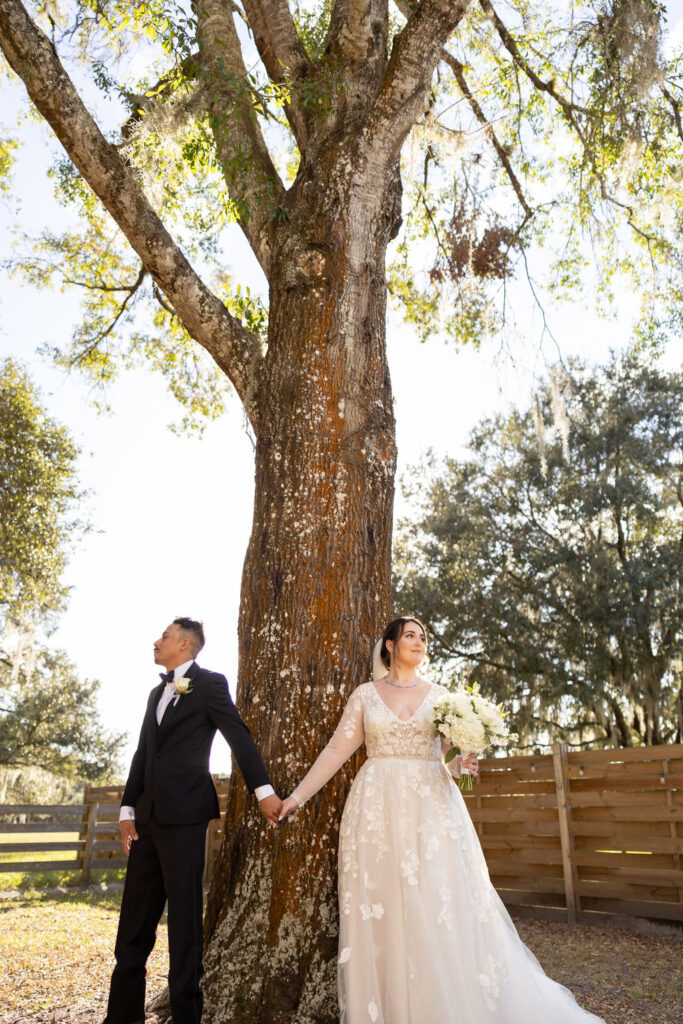 Newly wedded couple share a first touch moment. They stand beside a tall oak tree facing in opposite directions while holding hands before their outdoor ceremony at the Heart D Farm Wedding Venue.