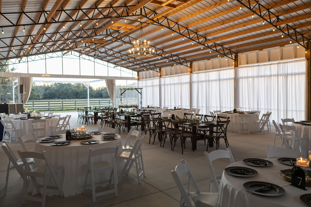 Inside the big barn pavilion wedding reception space. The ceiling is vaulted with black metal trusses, market lighting and a rustic chandelier. The white drapes are drawn closed on the side wall to tame the bright light coming through the clear vinyl doors. The end of the barn is open to the outside dance floor and has the gable end and partial walls are closed in with clear vinyl. The room is set for 10 guests with round tables with white garden chairs and wood farm tables with cross back chairs. The round tables are draped in white table linens while the farm tables are just the natural stained wood. The farm tables run through the center of the room while the round tables are set around the outside perimeter. It is a beautiful room with all the tables set with black chargers, wood rounds and floating candles in clear glass cylinders.