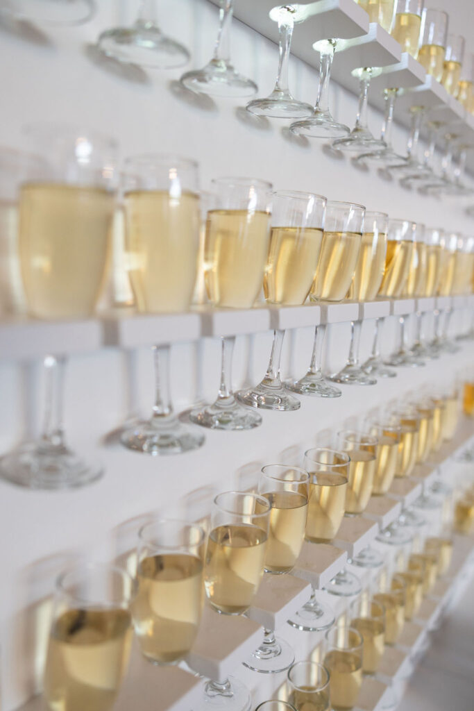 This photo is a close up of a white Champagne Wall filled with 96 clear glasses holding gold bubbly champagne waiting for the guests to arrive momentarily and lift a glass of their own for a champagne toast to the wedded couple!