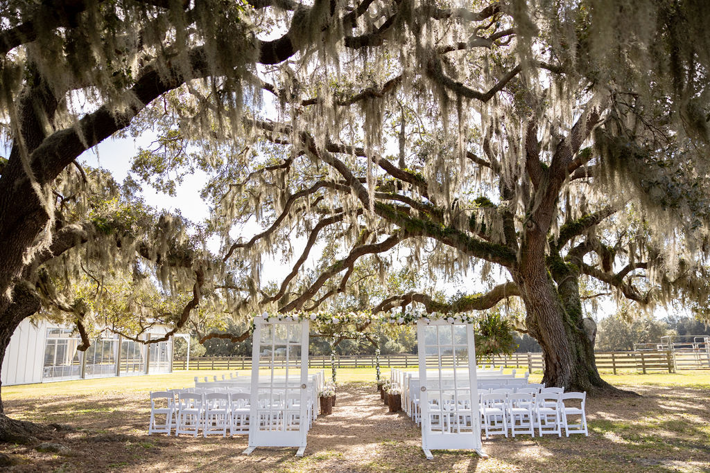 Beautiful outdoor ceremony site under the large Live Oak trees with draping moss and long limbs. The white church pews and white garden chairs are ready for the 100 guests. The white French doors with overhead decorative flowers frame the entry to the aisle and the traditional wood arch is at the front of the aisle awaiting the bride and groom and is decorated with greenery and white flowers.. The aisle is lined with four tree chunks on each side and each is decorated with greenery and white flowers. In the distance is four board fence and an open field. It is a gorgeous natural setting.