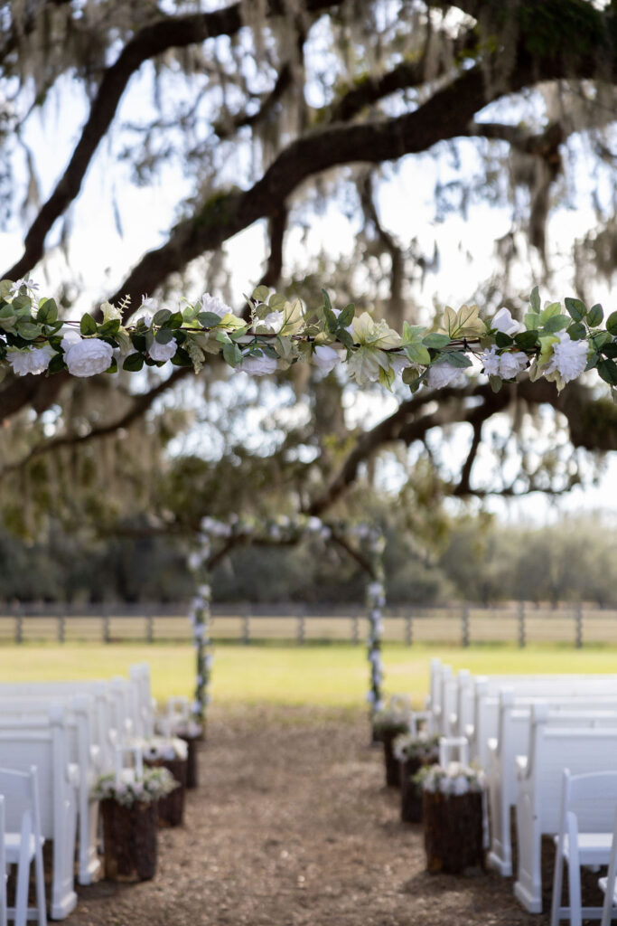 Outdoor wedding ceremony site set up under large ancient oak trees. The wedding arch is trimmed in greenery and white flowers. There are white church pews and white garden chairs awaiting the guests. The aisle is lined with chunks from trees and are decorated with greenery and white flowers. In he distance is a pasture with a four board fence. It is a lovely setting!