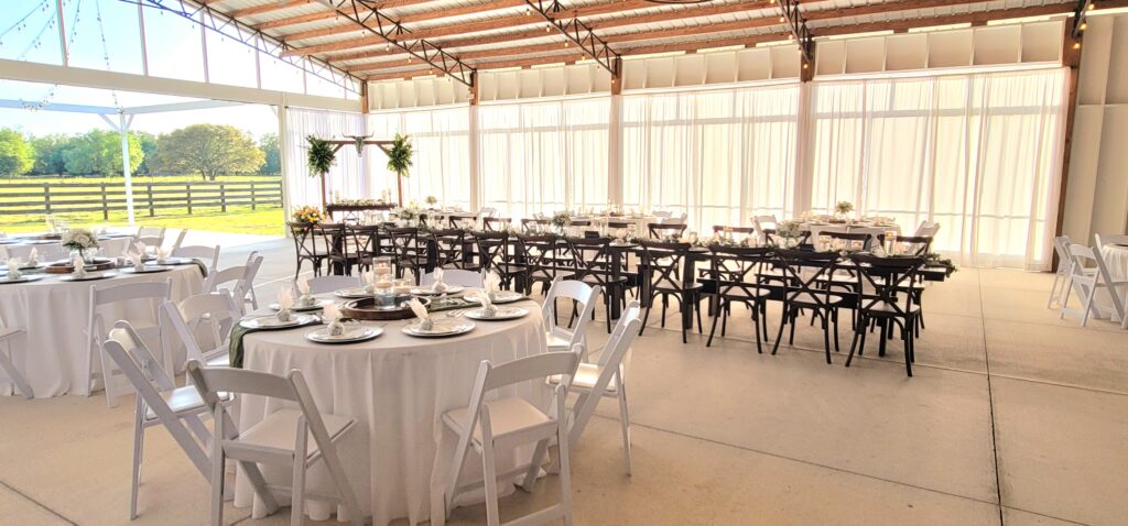 Gorgeous wedding reception area is set up for 100 guests.  The round tables are draped with floor length white linens. The wood farm tables make a lovely contrast against the closed elegant white drapes.  The couple decorated their traditional wedding arch with a cow skull and Florida greenery.  Elegance meets rustic ranch romance. 