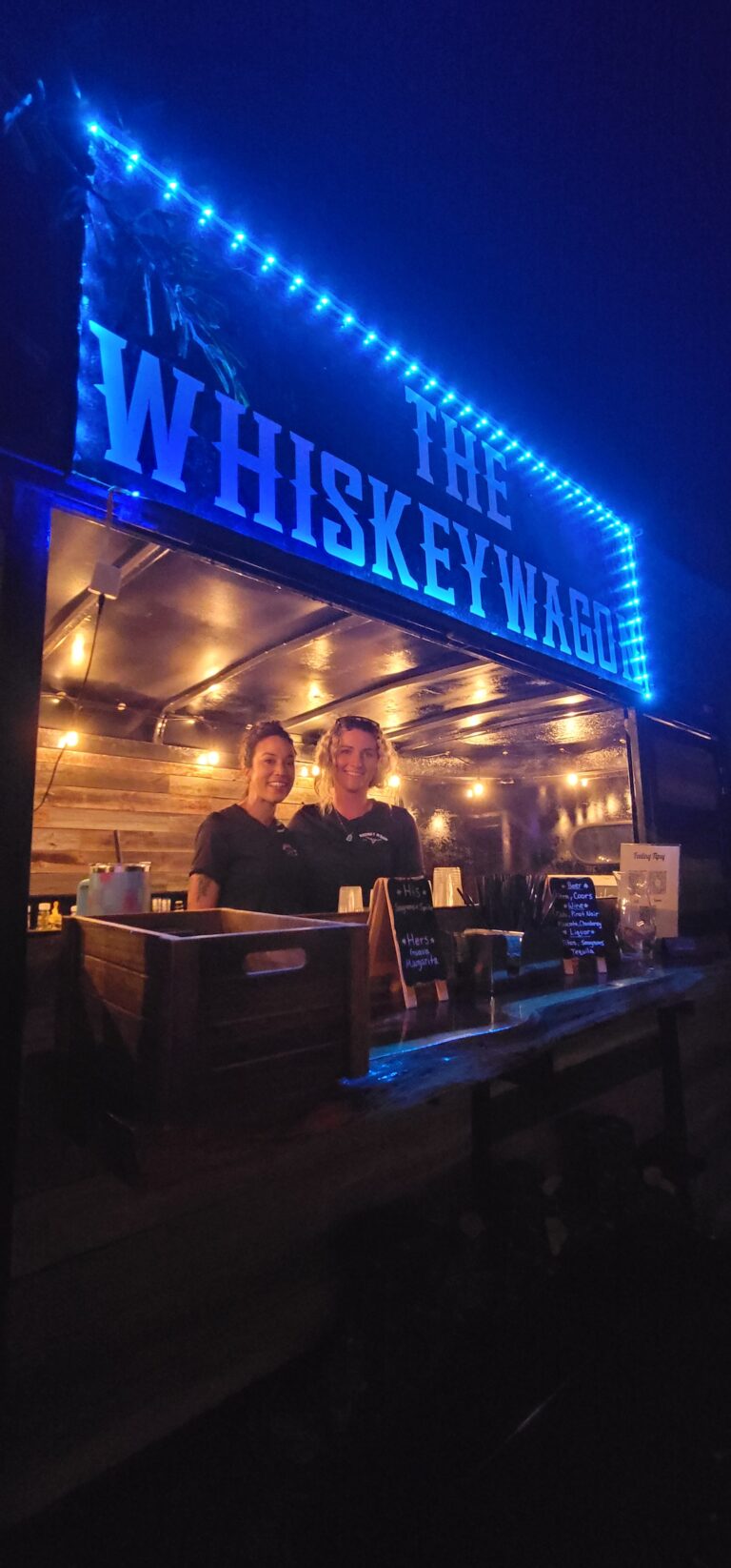 The Whiskey Wagon is a popular vendor at the Heart D Farm. The neon sign turns colors and is bright blue in the photo while the bartenders stand inside the horse trailer bar with smiles waiting to serve the wedding guests.