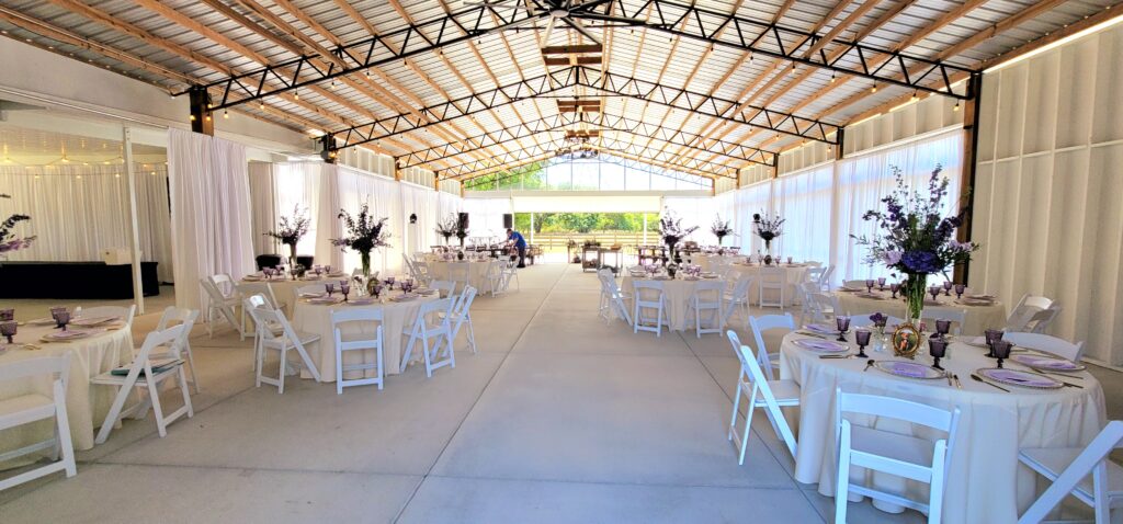 Beautiful wedding reception is set up and ready inside the glass wedding barn.  The elegant white drapes are closed to surprise the wedding guests when cocktail hour ends and the reception begins. The tall lavender and purple flower centerpieces are stunning under the high ceiling of the room.