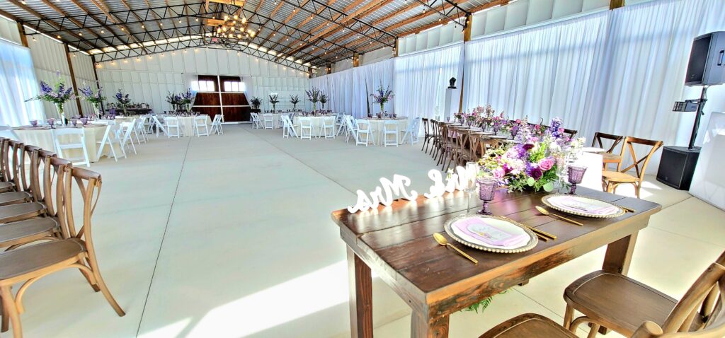 The Big Wedding Barn Pavilion is set with the dance floor inside for this newly wedded couple.  The sweetheart table is adorned with "Mr & Mrs" and lovely chargers and deep purple water goblets. The round tables are draped with floor length ivory tablecloths and the wood farm tables are placed to frame the dance floor. The tall lavender and purple centerpieces grace the center of the round tables and make a beautiful statement in the tall room.  The white drapes are closed to offer an elegant backdrop.