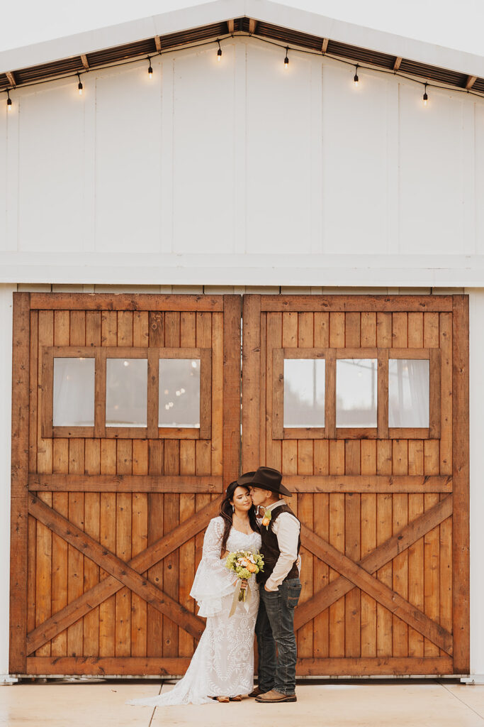 A newly wedded couple stand in front of the large wood barn doors. He is kissing her cheek and she is leaning into him as they share a sweet moment.