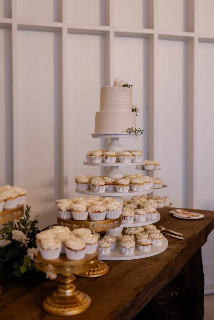 Beautiful small white wedding cake sits atop the cake display with dozens of white cupcakes on several levels and on separate cake stands.